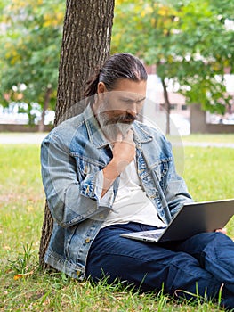 Serious middle aged man with laptop in city park. He sitting on the grass in the park under the tree. Vertical photo.