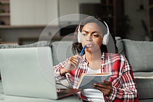 Serious millennial african american woman in wireless headphones studying with notepad and laptop