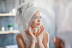 Serious millennial lady in bath towel looking at her face in mirror at bathroom