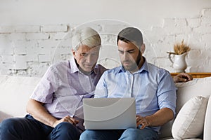 Serious millennial man helps to older father with laptop usage