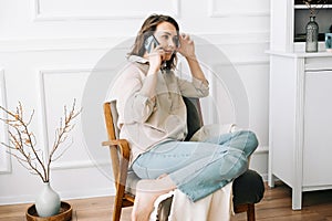 Serious millennial woman talks on the phone, sitting on a chair and looking out the window in her free time. Serious