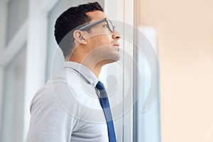 Serious mixed race businessman looking out of a window standing alone in an office at work. One hispanic standing and