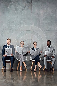 Serious multiethnic businesspeople sitting on chairs and reading newspapers in
