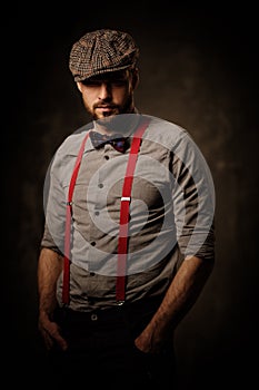 Serious old-fashioned man in tweed hat wearing suspenders and bow tie, posing on dark background.