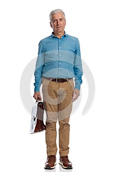 Serious old guy with bag wearing denim shirt and chino pants and standing