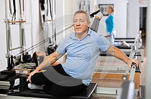 Serious old male in sportswear training twist rotation while using Pilates performer bed in gym