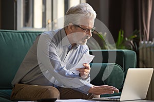 Serious old man holding paper paying bills online using laptop