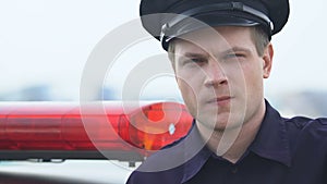 Serious patrolman in hat standing near squad car and looking to camera, enforcer