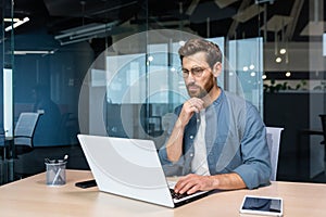 Serious pensive businessman in shirt thinking about decision sitting at table in modern office, man with beard is using