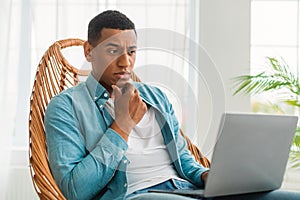 Serious pensive handsome young black man in casual looks at laptop, sits in chair in bright room interior