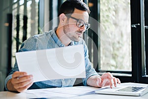 Serious pensive thoughtful focused young casual business accountant bookkeeper in office looking at and working with laptop and