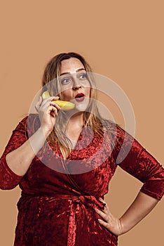 Serious phone call. Studio shot of shocked plus size woman in red velour dress holding banana near her ear and