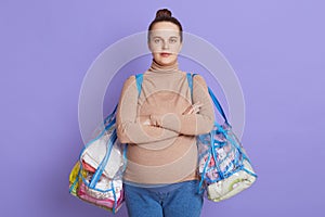 Serious pregnant woman with arms crossed on her chest, looks at camera, dresses beige sweater and jeans for future mother, holding