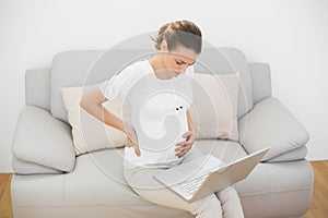 Serious pregnant woman touching her injured back using her notebook