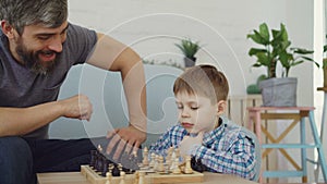 Serious preschool child is playing chess with his parent thinking about next move and moving chesspieces while his