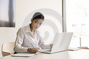 Serious pretty Indian business professional girl writing notes at laptop