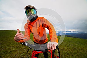 Serious professional cyclist in sportswear and helmet standing with a downhill bike on top of a hill, showing thumbs up