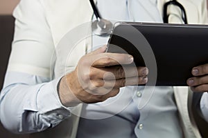 Serious professional doctor in white coat and stethoscope holding modern touch screen gadget using digital tablet computer at work