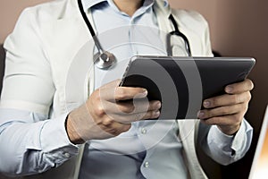 Serious professional doctor in white coat and stethoscope holding modern touch screen gadget using digital tablet computer at work