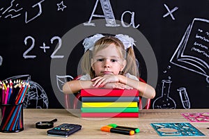 Serious and puzzled schoolgirl sitting at the desk with a pile of books under the chin, surrounded with school supplies