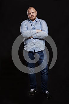 A serious red fat man with a beard is standing with his arms crossed on his chest. Full height. Black background. Vertical