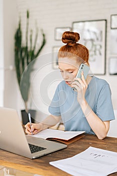 Serious redhead young businesswoman is working on laptop computer and talking on cell phone