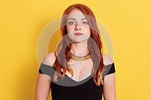 Serious sad ginger female, looking directly ar camera, woman with red lips wearing black elegant dress and golden necklace, posing