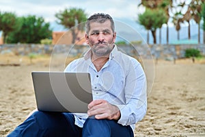 Serious sad mature man with laptop sitting on cloudy beach.