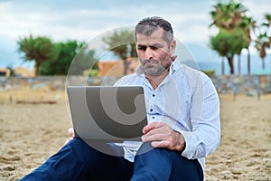 Serious sad mature man with laptop sitting on cloudy beach.