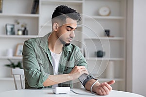 Serious sad young middle eastern man with beard measures blood pressure with tonometer, feels bad photo