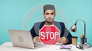 Serious self confident office worker in business suit showing stop sign sitting at laptop on his workplace, stop work harassment