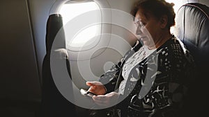 Serious senior airplane passenger woman typing messages on smartphone messenger app sitting on airplane window seat.