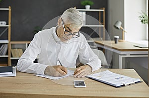 Serious senior businessman sitting at office desk and taking notes in his notebook