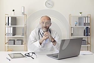 Serious senior doctor looking at the camera while sitting at a with laptop in a medical clinic office.