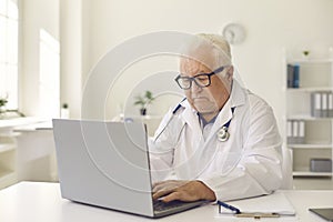 Serious senior doctor typing on laptop keyboard sitting at desk in clinic office