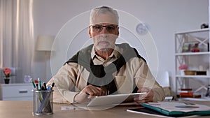 Serious senior man holding tablet and looking at camera, planning budget app