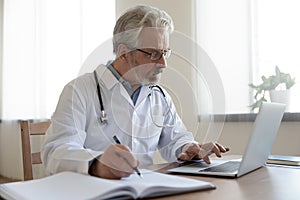 Serious senior physician using computer app making notes in journal