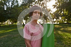 Serious senior woman with fitness mat in park