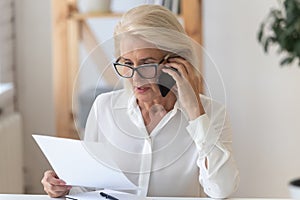 Serious senior woman talk on cellphone discussing paperwork