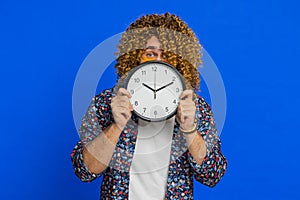 Serious strict stylish man holding office clock hiding behind checking time on watch obscuring face