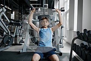 Serious strong teenager boy training with dumbbells while sitting on bench