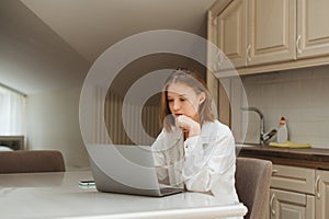 Serious student girl sitting at a table in the kitchen and studying, looking at a laptop screen.Attractive freelance girl working