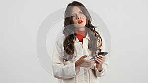 Serious stylish girl annoyedly looking in camera using smartphone over white background