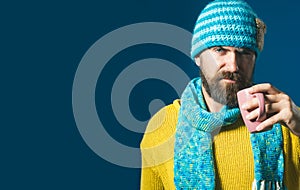 Serious stylish man in knitted sweater and hat wrapped in scarf with mug of coffee or tea in cafe. Handsome bearded man