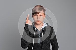 Serious Teen boy with raised index finger, looks at camera, stands over grey background, wearing in casual clothes