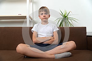 Serious teen boy sitting at home on sofa, legs and arms crossed