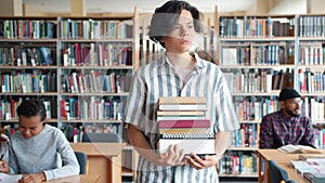 Serious teenage boy walking in library with bunch of books looking around