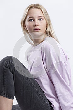 A serious teenage girl with freckles is sitting. Beautiful blonde in a pink jacket and black jeans. White background. Vertical