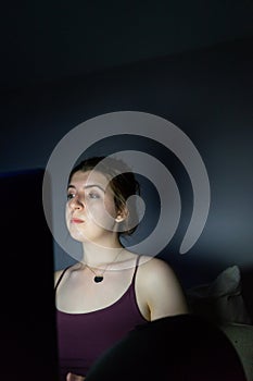 Serious teenager girl sitting near laptop in dark room. Personal computer and woman. Working at night from home in the living room