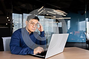 Serious thinking asian businessman working inside modern office, mature man in shirt and glasses using laptop at work